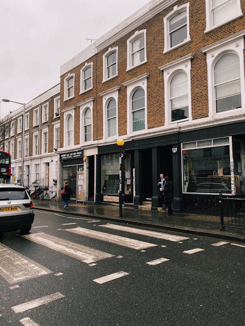 The image depicts a busy street scene in Kensington, showing a three-storey brick building with large ground-floor windows and white decorative window arches on the upper floors. The pavement is wet, 