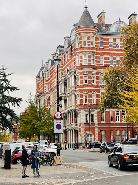 The image depicts a busy street scene in Kensington, showing a three-storey brick building with large ground-floor windows and white decorative window arches on the upper floors. The pavement is wet, 