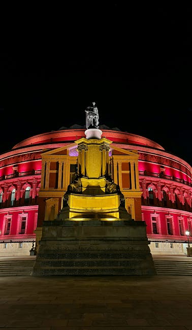 An exterior view of a large, circular, historic building with a domed roof and ornate architectural details, located near Royal Albert Hall. In the foreground, a stone monument with multiple statues a