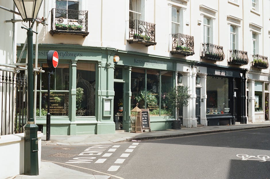 Exterior view of a row of mixed-use buildings on a street in Kensington, featuring a ground-floor flower shop with large glass windows displaying floral arrangements, a blackboard sign outside advertising flowers, and a small potted plant near the entrance. The upper floors have white-painted façades with ornate black iron balconies adorned with potted plants and flowers. A green and white building with floral shop signage is adjacent to a neighboring property with a black storefront. The street includes a pedestrian crossing with white dashed lines and a bicycle lane, along with a no-entry road sign mounted on a pole near the corner. The scene is well-lit with natural daylight, and there are no people visible in the image. The overall setting depicts a quiet, urban residential and commercial area suitable for house removals and furniture transport services by Man With a Van Kensington, highlighting typical elements involved in home relocation logistics.